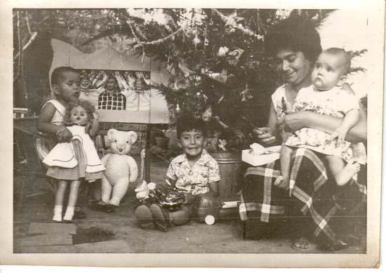 Fotografía en escala de grises en que se aprecian una niña, un niño y un bebe en los brazos de un adulto. LA escena se contextualiza en un arbol de navidad y los niños tienen en sus manos juguetes como una muñeca y un peluche. 