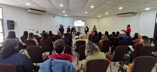 personas sentadas en un salón escuchan a los expositores que presentan en el escenario