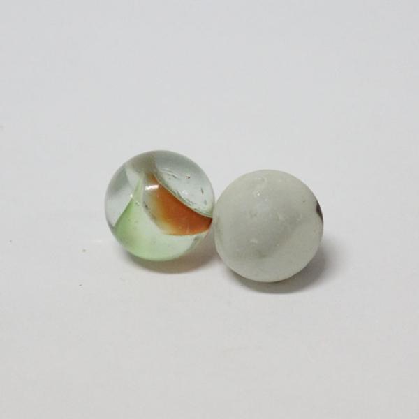 Imagen de fondo gris, en el centra de aprecian dos canicas o bolitas. La primera es de vidrio y en su interior se ven unas líneas verdes y naranja. A su costado hay otra bolita de color blanco.