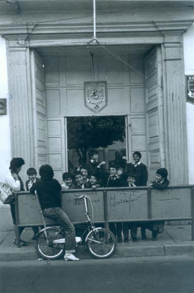 Fotografía monocromatica del ingreso de una escuela urbana, en el centro iun grupo de niños de distintas edades apoyados en una barrera metalica.