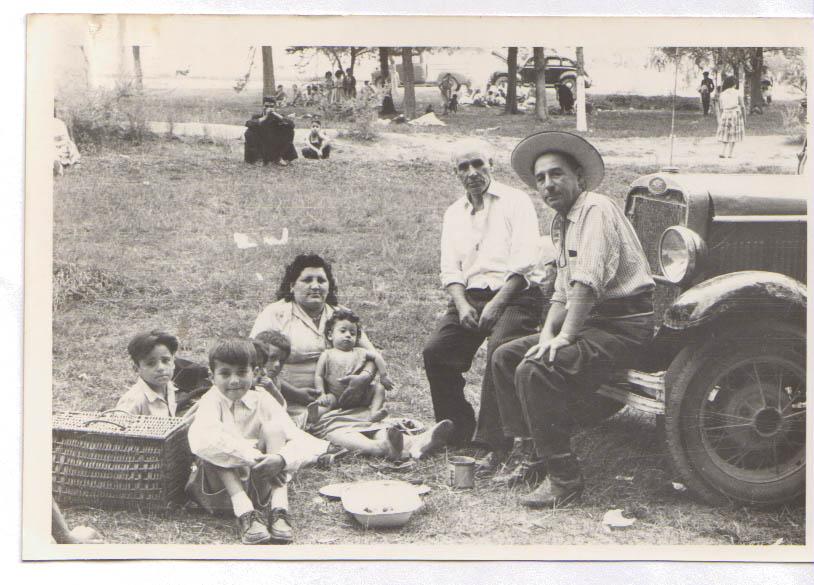 Fotografía monocromática, compuesta de una escena familiar de 7 integrantes que disfrutan un día de picnic.Sentados en el capo de un automóvil, un anciano y un hombre adulto. En el suelo, sentado en el cesped tres niños de edades similares, una mujer con un bebé en sus brazos.