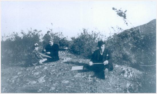 Fotografía monocromática que ilustra una escena en el cerro, compuesta por dos adultos y una niña sentados en el suelo preparando sus volantines.