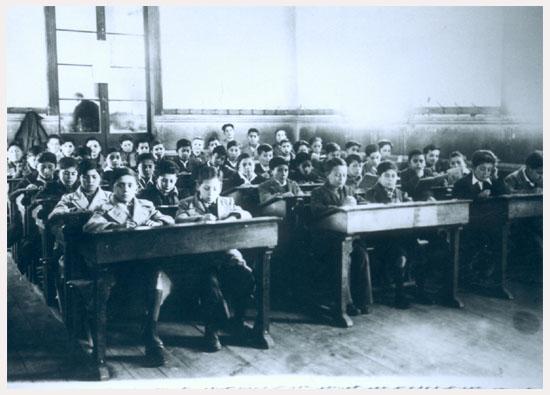 Fotografía monocromatica de una sala de clases en el Instituto O'Higgins de Rancagua. En ella se aprecia un grupo de niños sentados en sus pupitres dobles, la sala está distribuida en tres filas, en el fondo se observa una pared con una ventana de gran tamaño que aparentemente permite mirar hacia otra sala de clases.